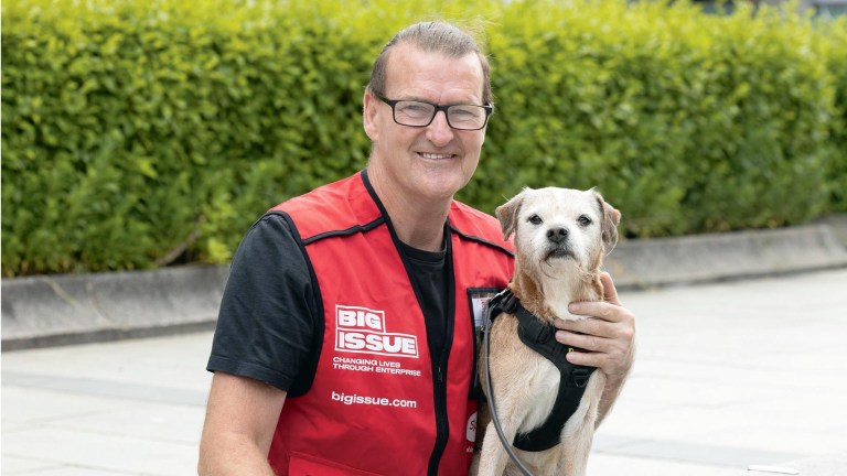 A smiling man wearing glasses and a red Big Issue vest stands on a sidewalk, holding a small tan dog. The man has short gray hair and is wearing a black shirt under his vest. The dog is looking directly at the camera. Behind them is a green hedge.