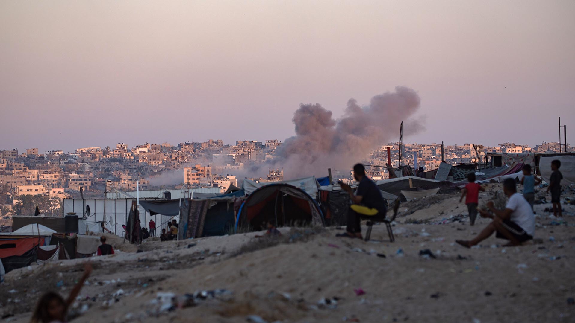 Smoke rises following an Israeli air strike as internally displaced Palestinians sit next to their tents in Khan Younis camp, southern Gaza Strip