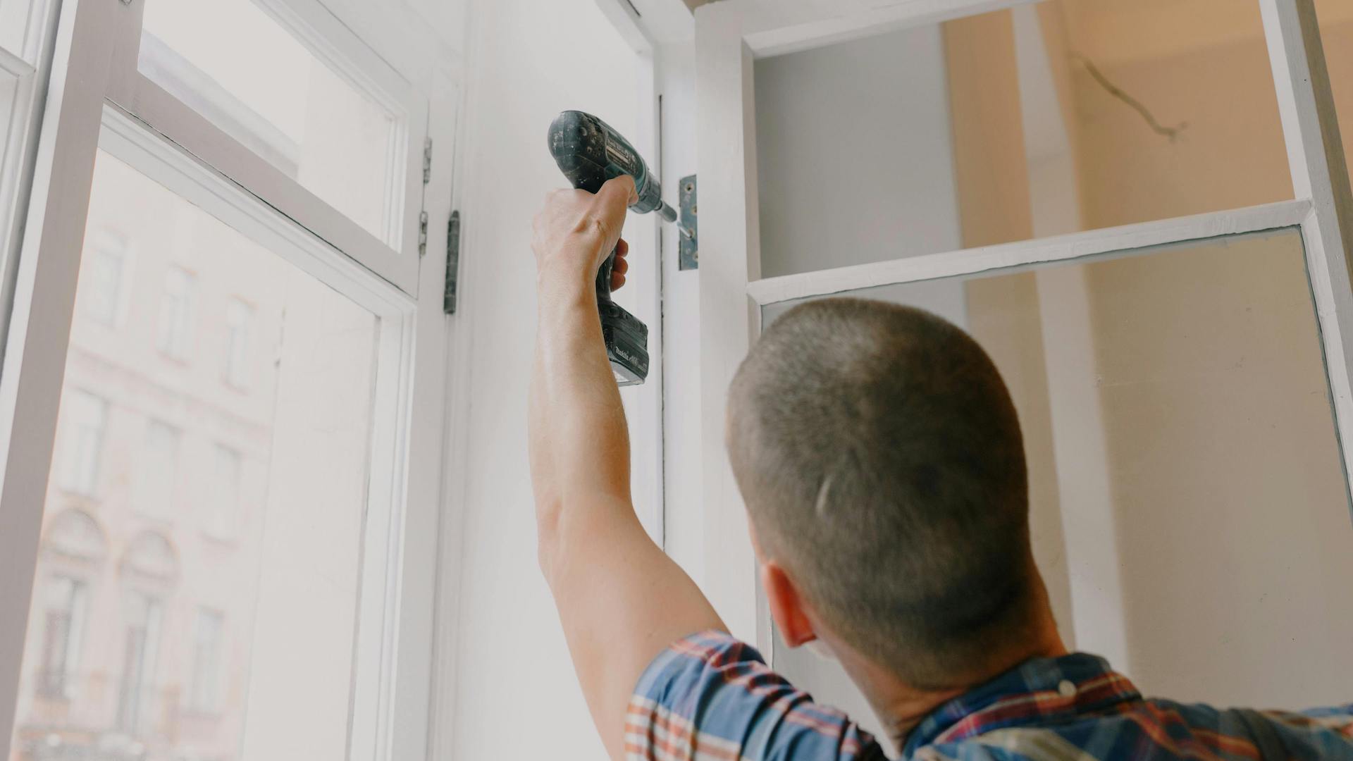 a man using an electric screwdriver to screw into a hinge on a window