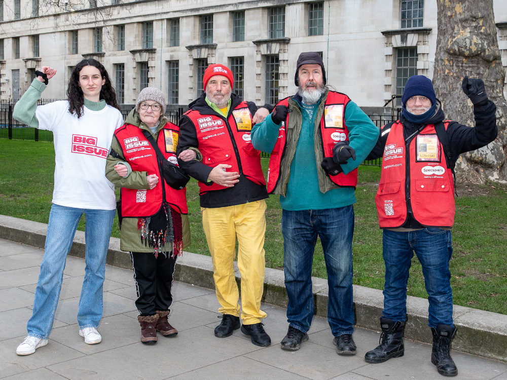 Group of 4 big issue vendors and staff member stand arm in arm, two of them have raised fists