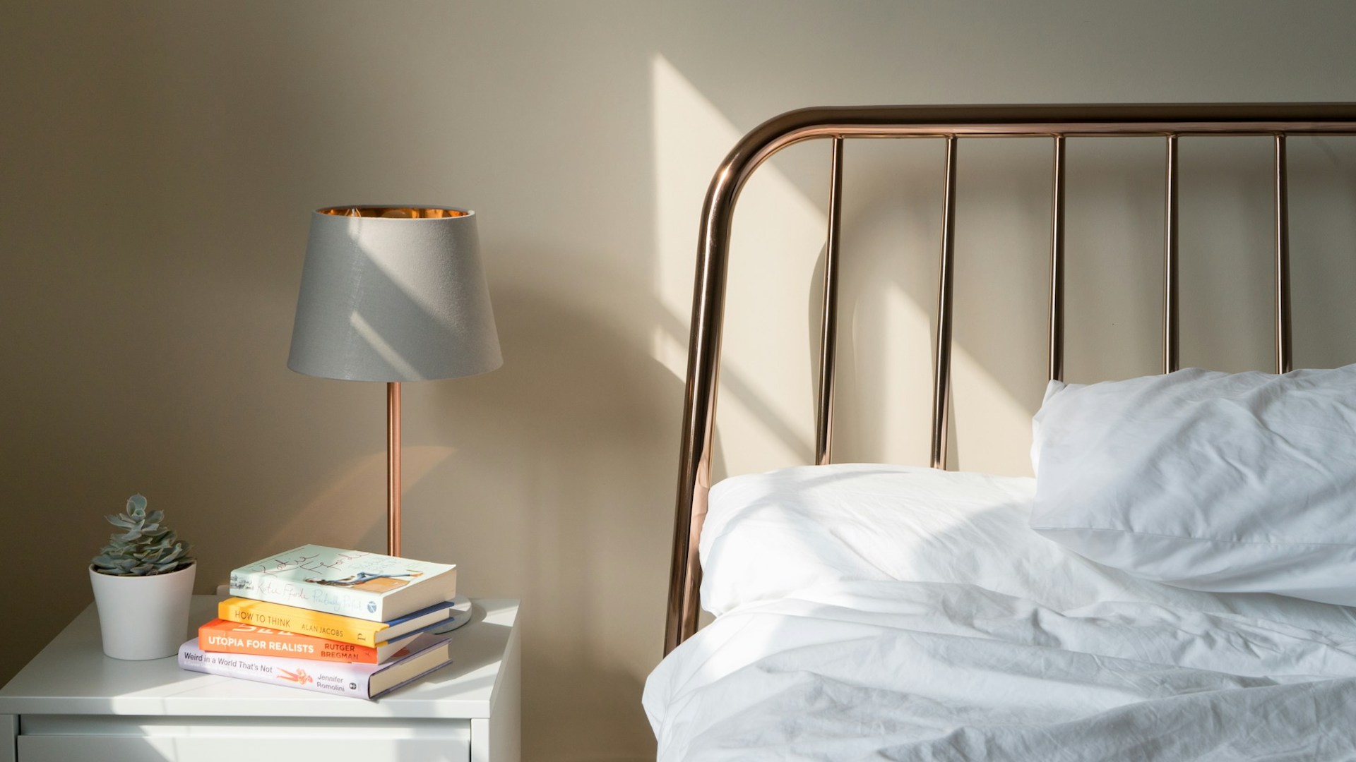 one side of a bedframe against a wall next to a bedside table with books and a plant on it