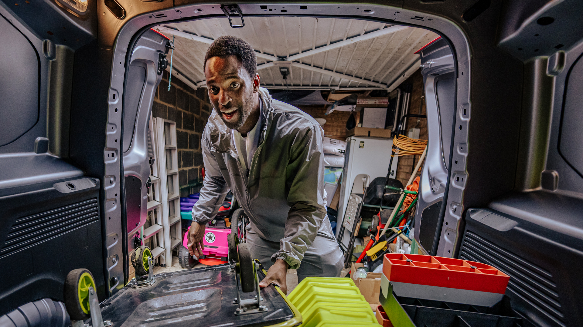 A smiling man wearing a light grey jacket leans into the back of a Citroën ë-Berlingo Van, surrounded by colourful fitness equipment and storage containers. The image is taken from inside the van looking out through the rear doors, showing the van's spacious cargo area and interior features.