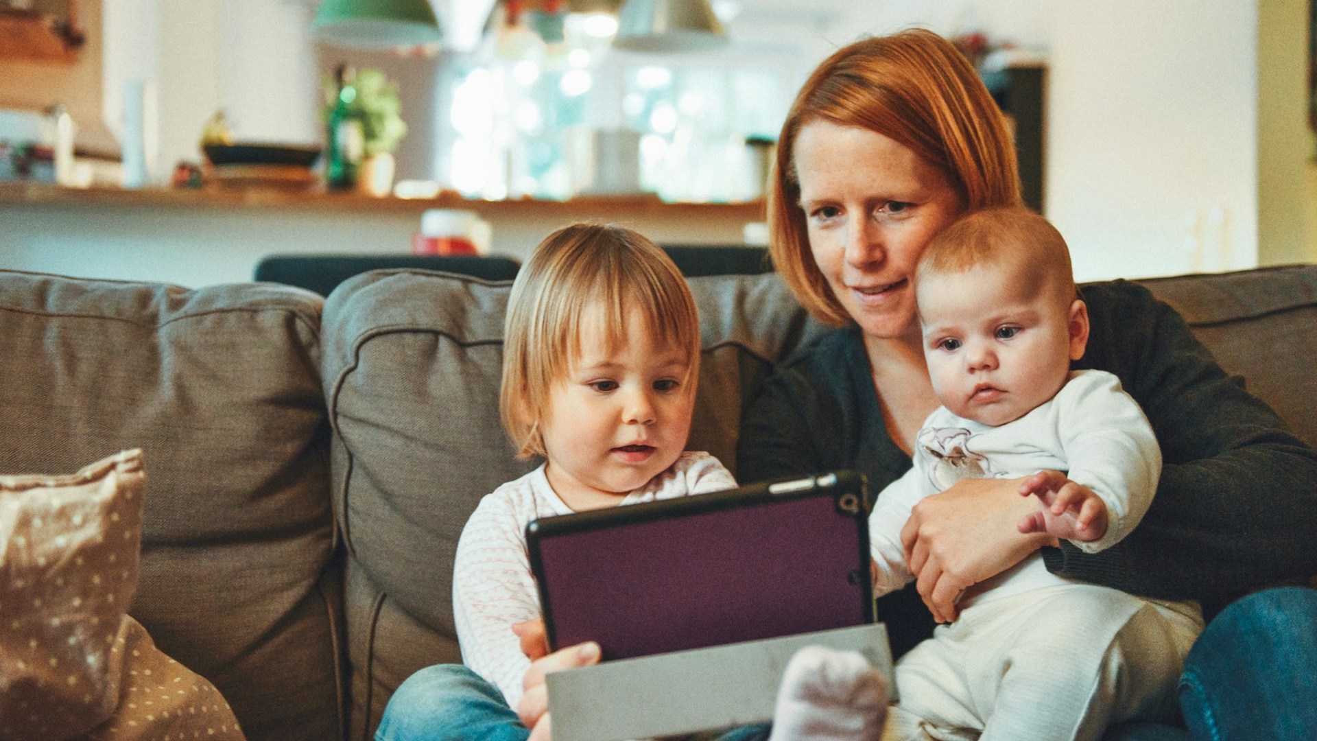 a woman sat on a couch with two young children on her lap, looking at an iPad