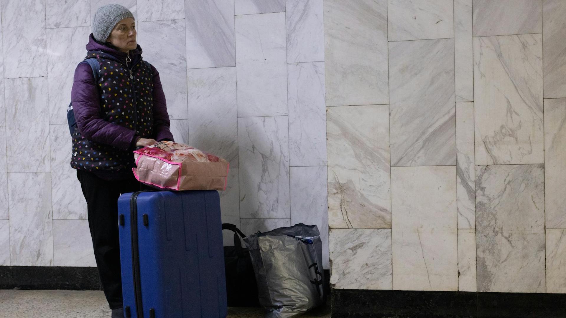 a woman wearing a coat and beanie hat stands with a suitcase and two large bags of belongings in front of a grey wall