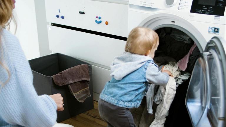 a crouching woman watches while a toddler pushes clothes into an open washing machine