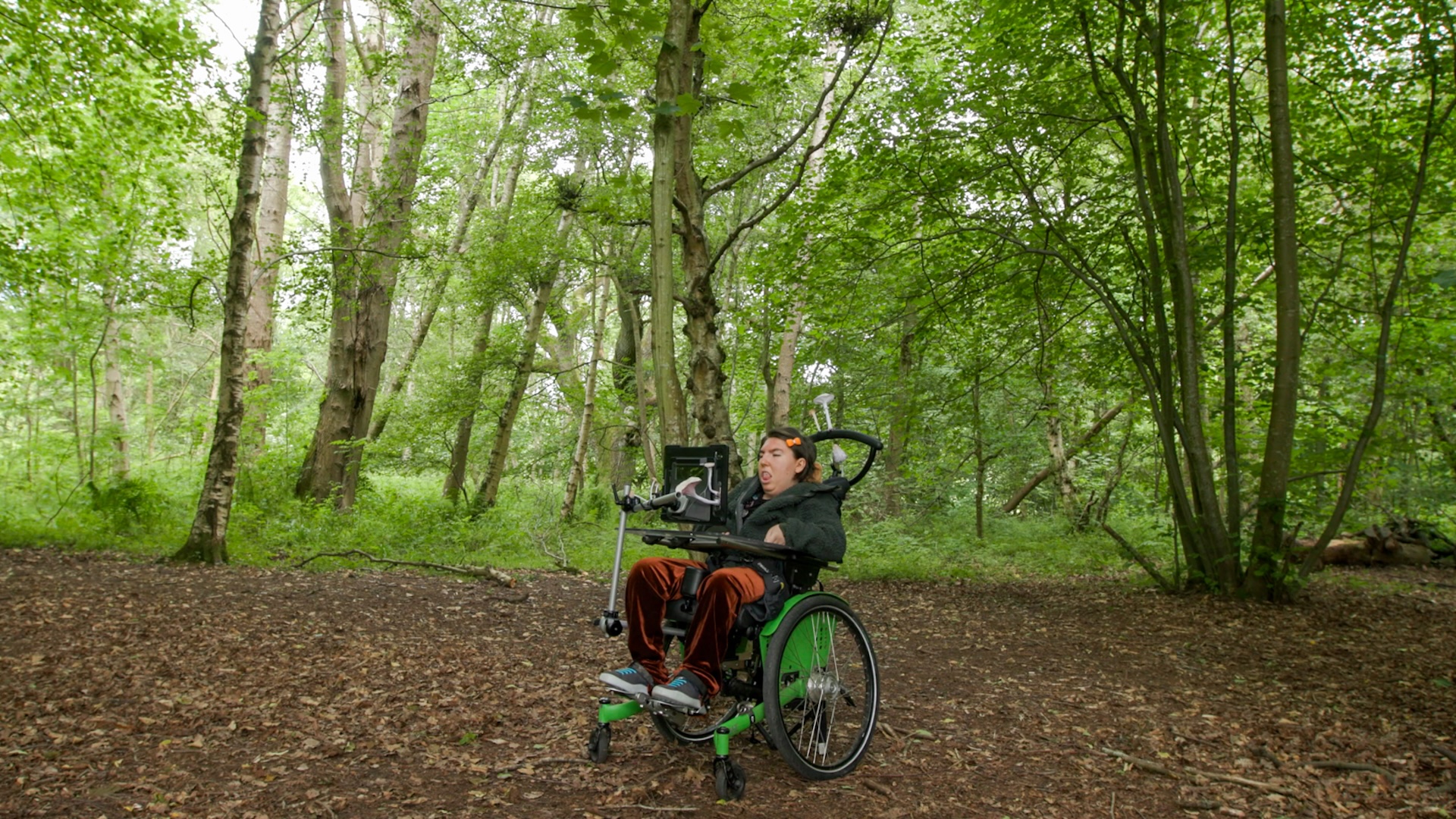 Greta, a white woman and wheelchair user, sits in the middle of a green forest on location during filming for A World Beneath Us. It is Bumpy, cool, warm, trees, plants, insects, staggering, quiet, light, dark (shade), tan, green, brown.