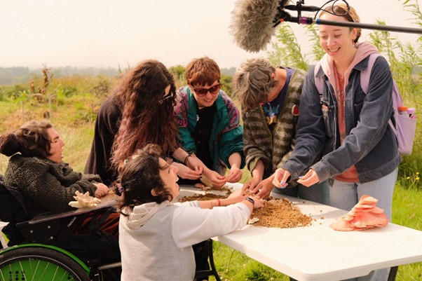 A group of women are outside standing or sitting around a table handling soil and mushrooms. They are smiling, laughing and concentrated on the feeling of the materials. The scene is funny, scared, smooth, mycelium, mushrooms, network, dusty, wet, warm, rainbow (of colours, clothes), pale blue, green, brown, yellow