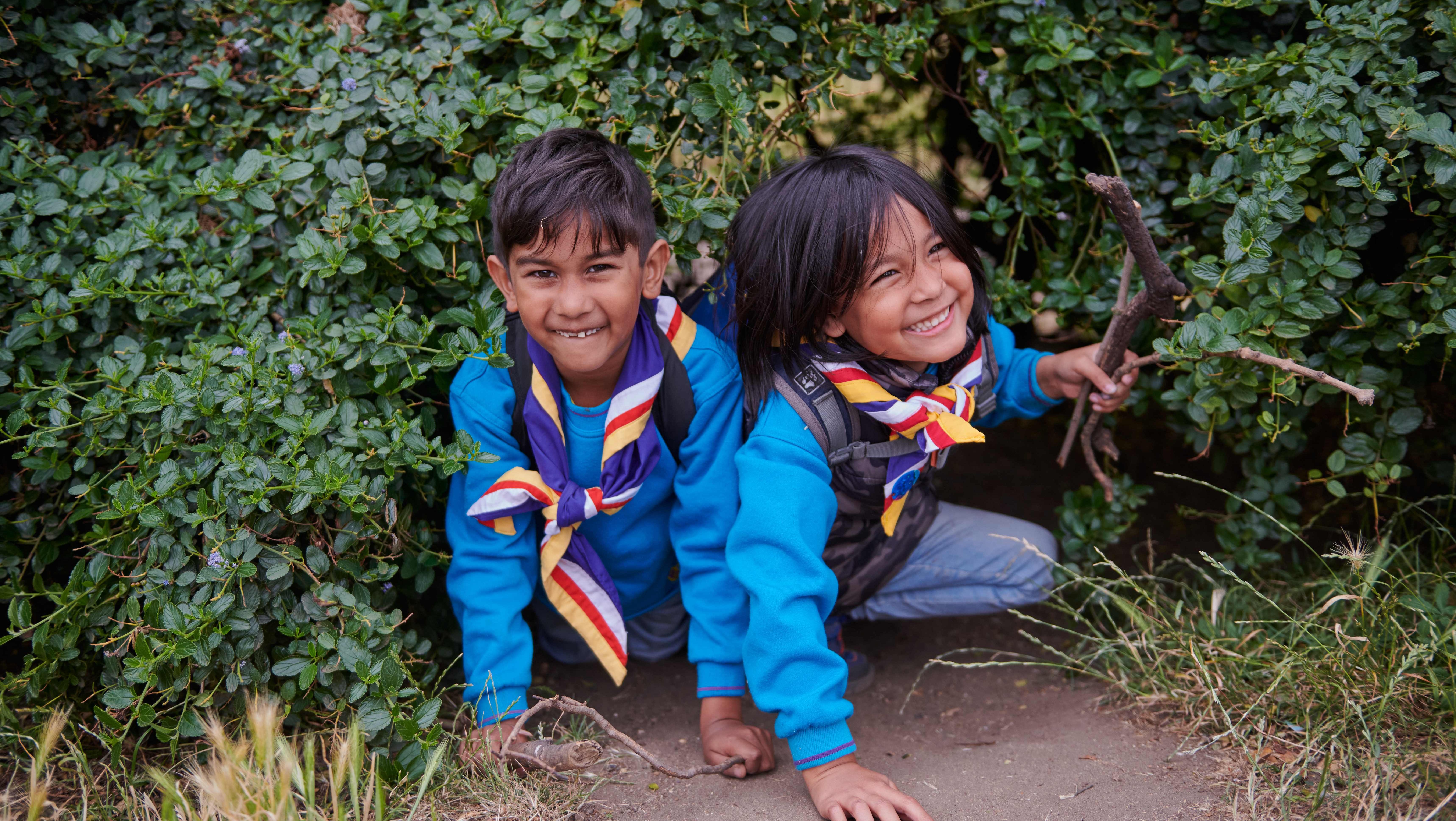 Two young children wearing beavers uniforms emerge from a hedge