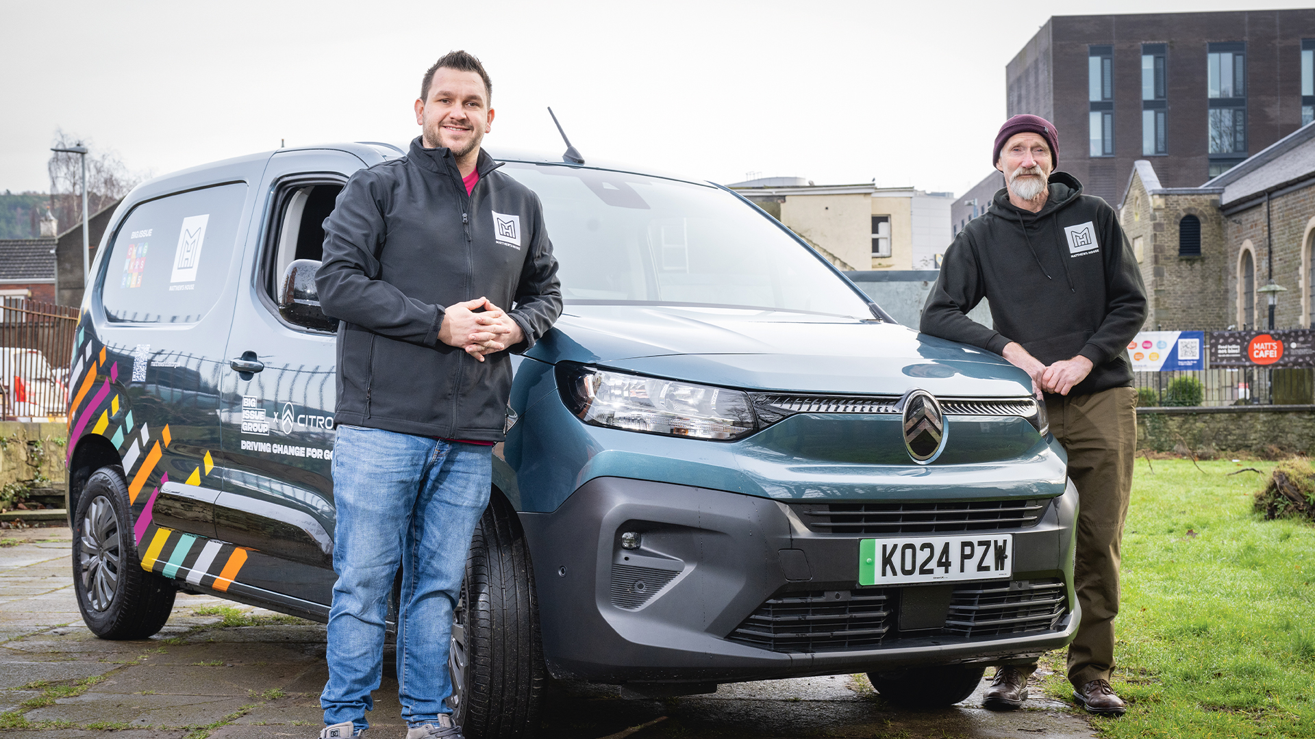 Two men stand in front of a Citroën electric van with a green number plate. The man on the left, wearing a black zip-up jacket with a white logo, blue jeans, and grey trainers, has short dark hair and a beard. The man on the right, wearing a black hoodie with a white logo, brown trousers, and brown shoes, has a white beard and a burgundy beanie. The van has a distinctive multicoloured design on the side, featuring diagonal stripes in orange, yellow, pink, and white. Logos for “Big Issue Group” and “Citroën” are visible on the van, along with the phrase “Driving Change for Good.” The background includes a mix of modern and older buildings, a church, and a grassy area.