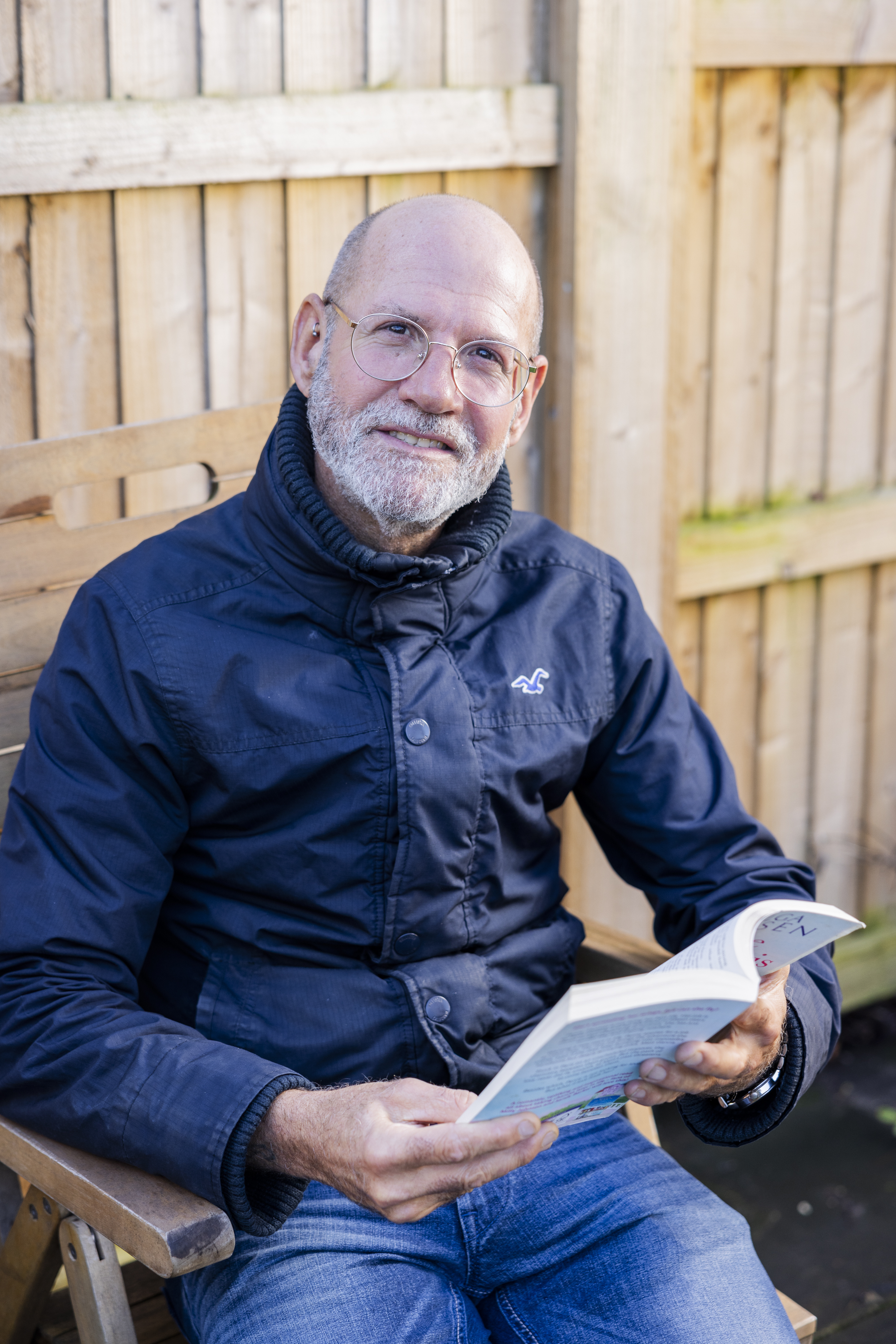 A man in a navy jacket sits on a wooden bench against a fence, reading a book and smiling at the camera. He wears round glasses and has a grey beard.