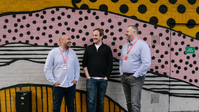 L-R: Big Issue job coach Shak Dean, Recruit candidate Kane, and Head of BIR operations Stuart Greenway stand in front of a wall in Finsbury Park, London, near Big Issue's head office.