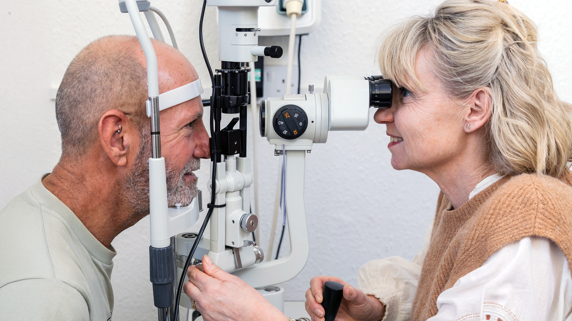 An optometrist and a person having an eye exam interact warmly during the examination, with the patient positioned at a standard optometry machine while the optometrist adjusts the equipment.