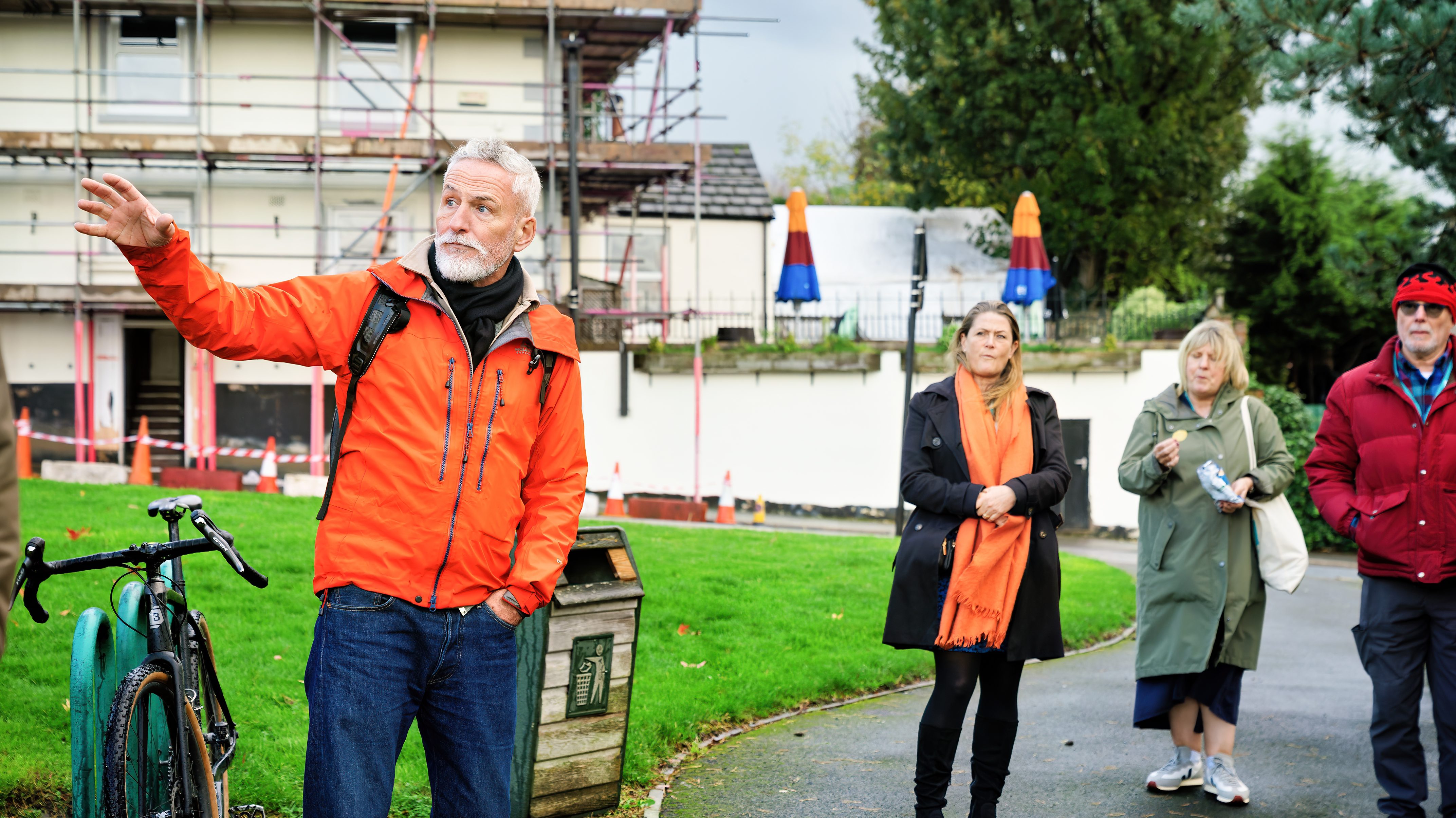 A man in an orange jacket stands with his arm out while three people look in the background